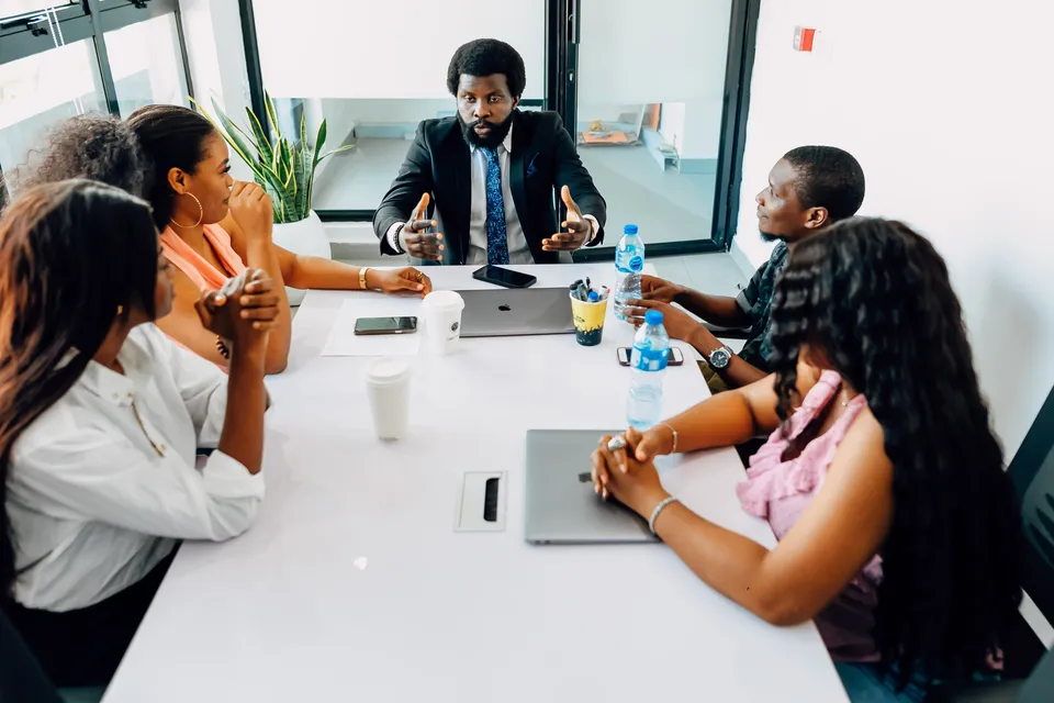 Five people around a white table in a bright meeting room, one of them speaking with open hands
