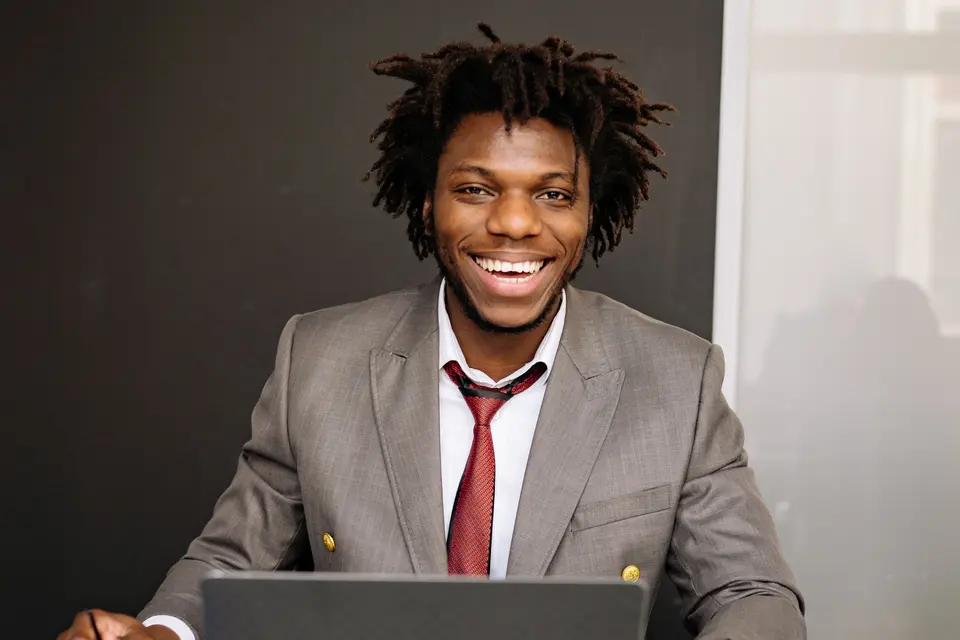 A man in a grey suit smiling at an office desk with a laptop in front of him