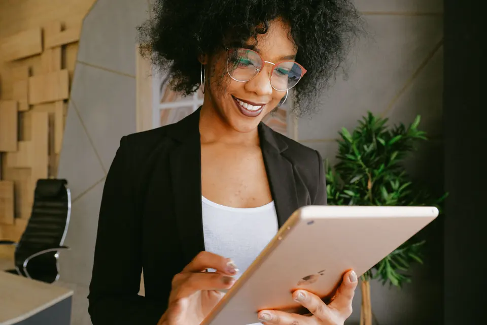 A woman in a black blazer smiling as she looks at a tablet in a bright office