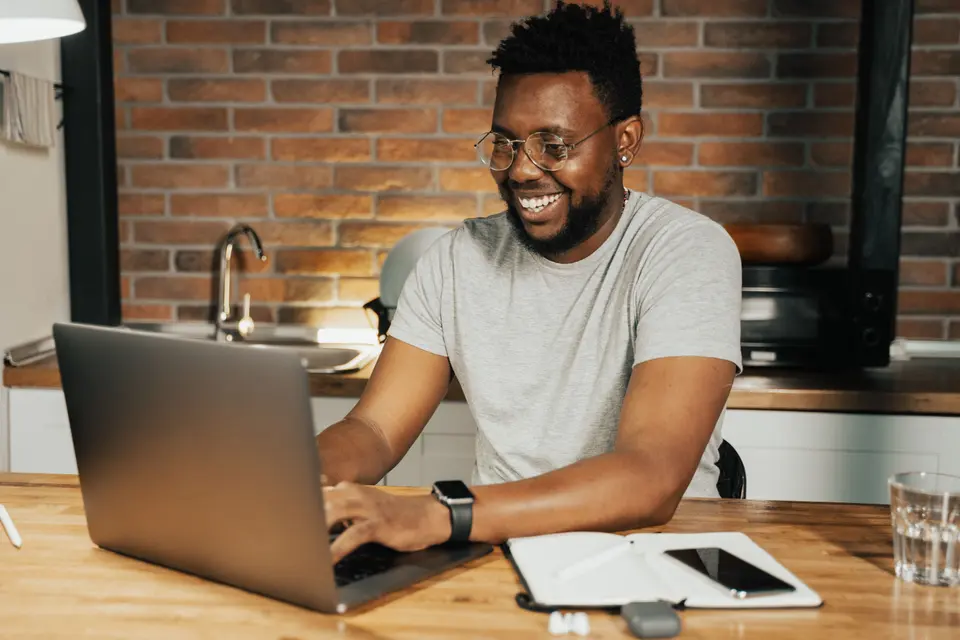 A man smiling as he works on a laptop at a wooden kitchen table, a phone and notebook beside him