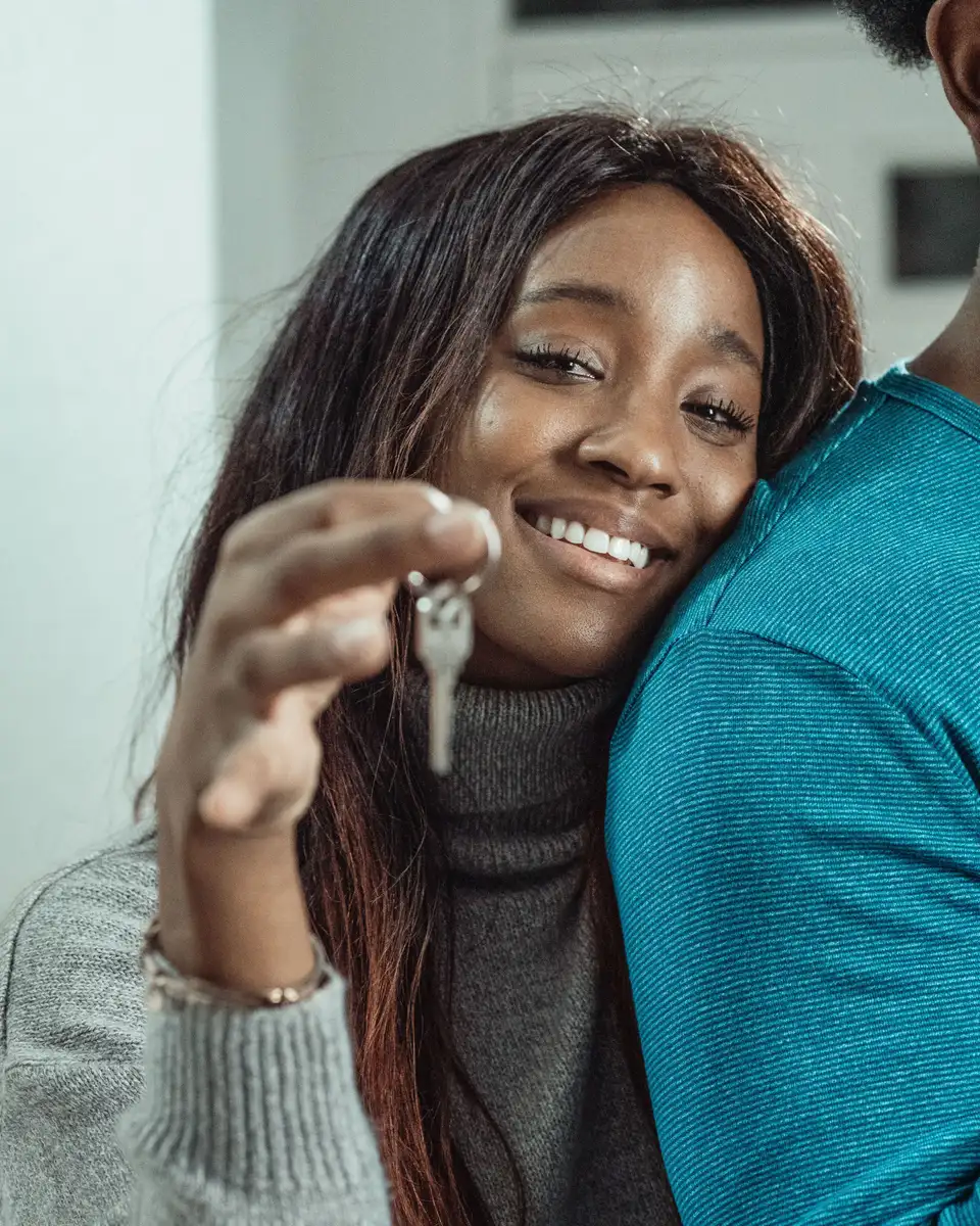 A smiling woman holding up a house key while a loved one hugs her at home