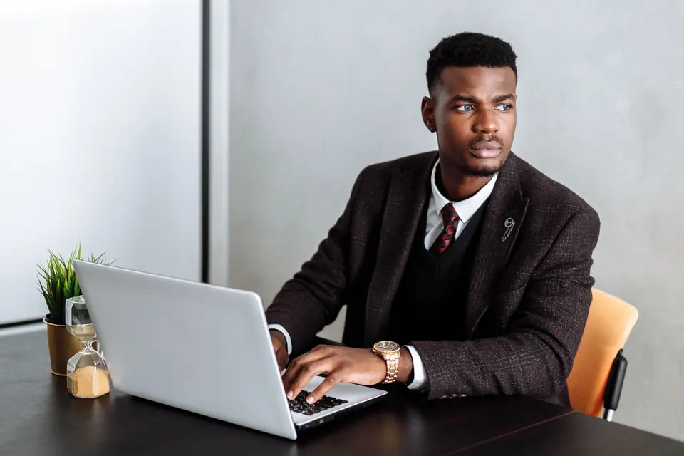A man in a dark jacket working on a laptop at a desk in a bright office
