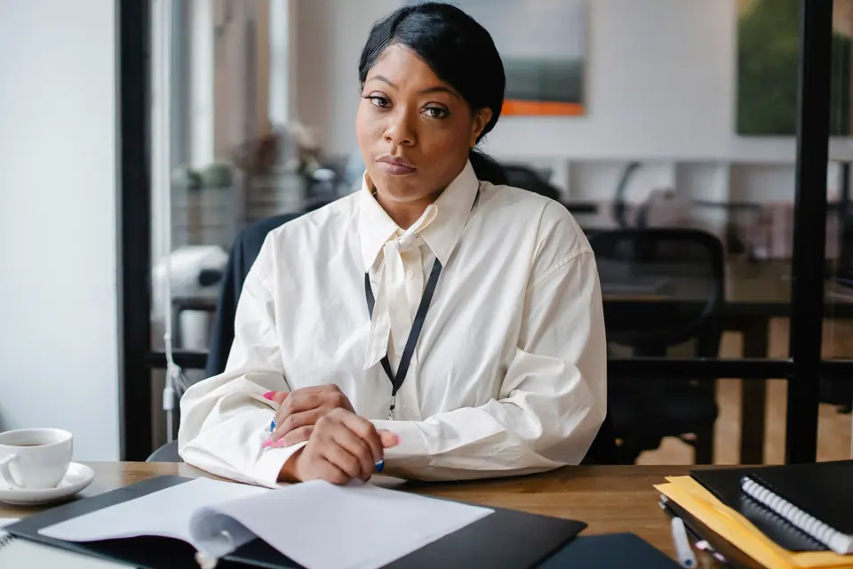 A woman in a white blouse sitting at a wooden desk with a folder of paperwork in front of her