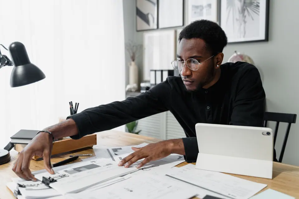 A man in round glasses and a black shirt leaning over a wooden desk of property plans, beside a tablet