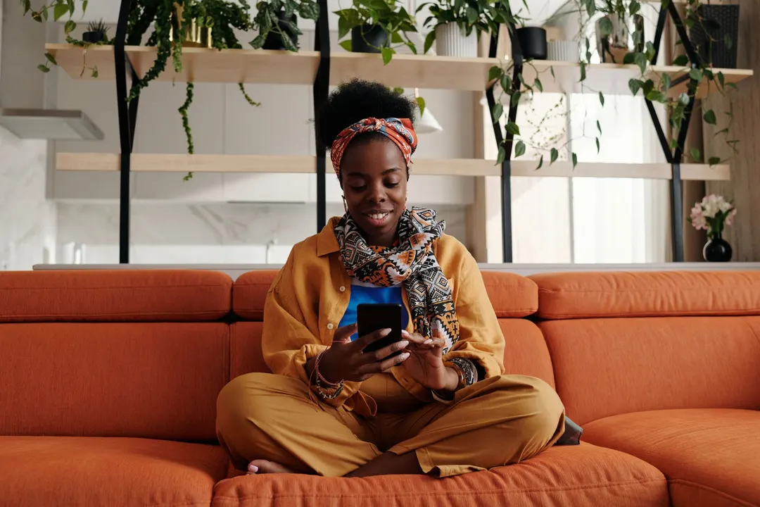 A woman smiling at her phone on an orange sofa in a bright, plant-filled living room