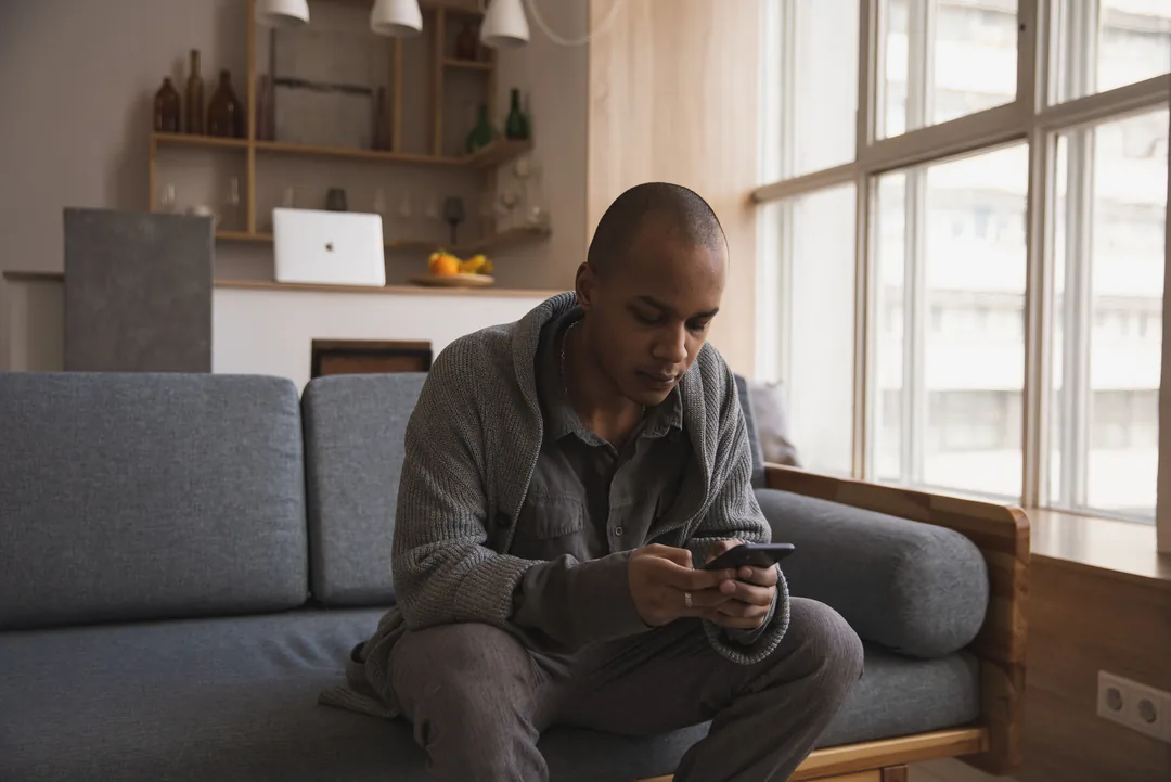 A man sitting on a grey sofa beside a large window, looking at his phone at home