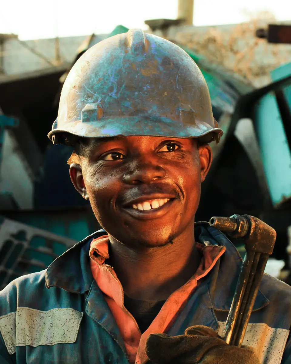 A smiling worker in a dusty hard hat and blue overalls holding a wrench against his shoulder