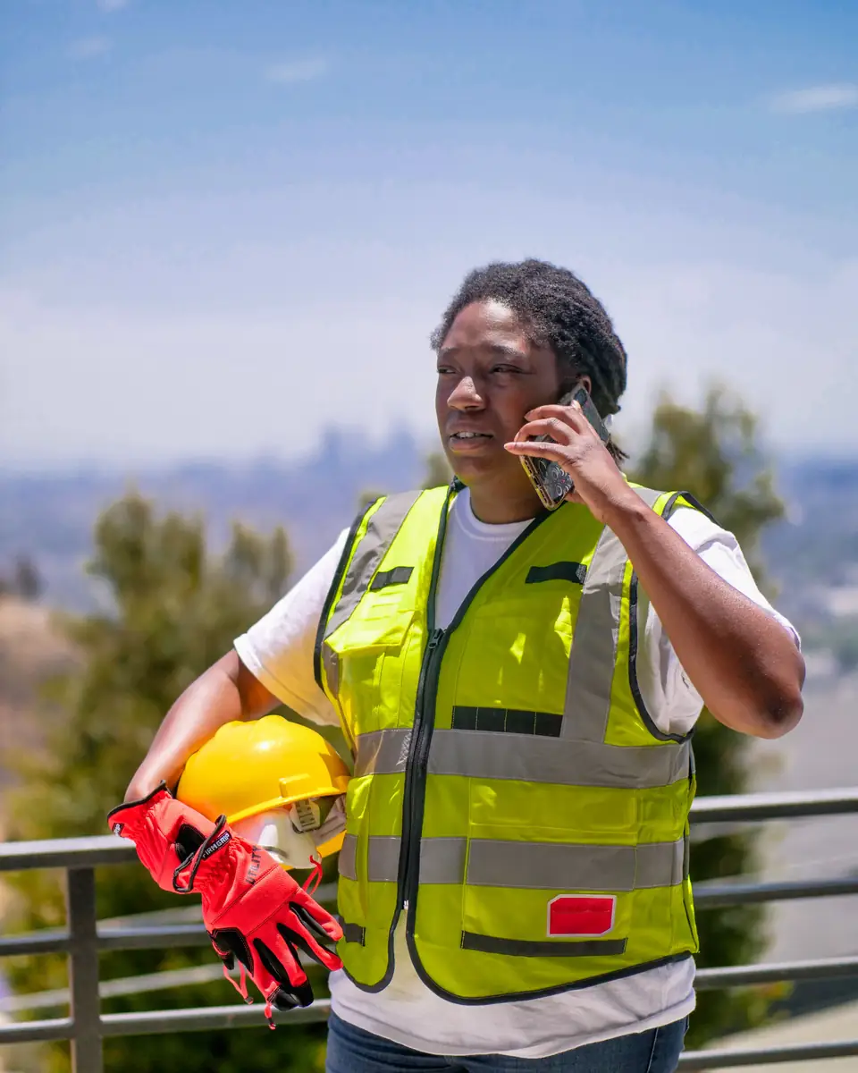 A contractor in a yellow safety vest on a phone call outdoors, a hard hat and red work gloves in her arm