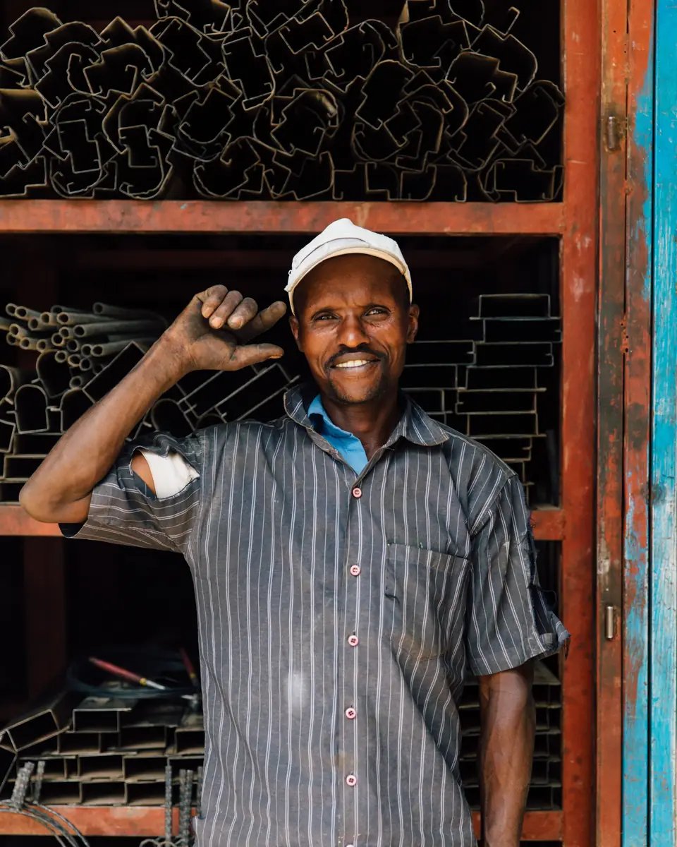A smiling metalworker in a cap and a striped work shirt, one hand raised, in the doorway of his workshop with steel profiles stacked behind him