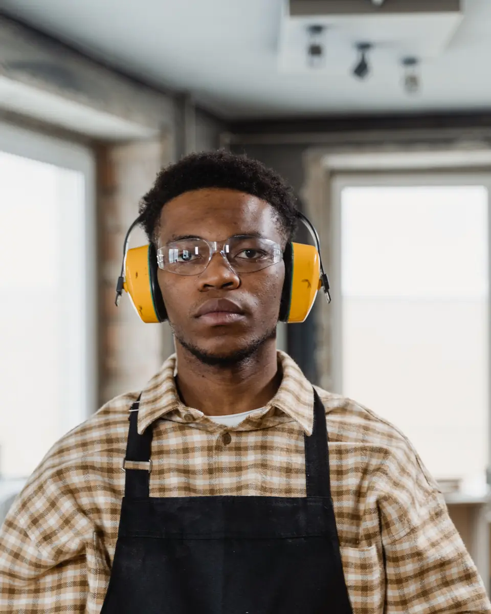 A young craftsman in yellow ear defenders, clear safety glasses and a black work apron standing in a bright room